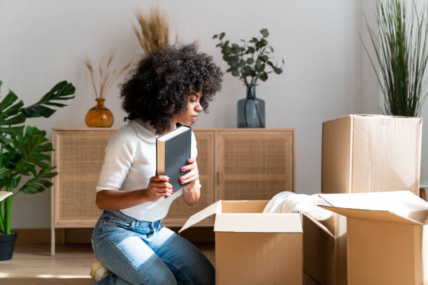 Beautiful woman unpacking her favorite books in her new apartment. Kneeling on the floor taking books out of cardboard boxes to place on the living room shelf.